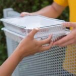 Close-up of two people exchanging aluminum food containers over a metallic fence outdoors.